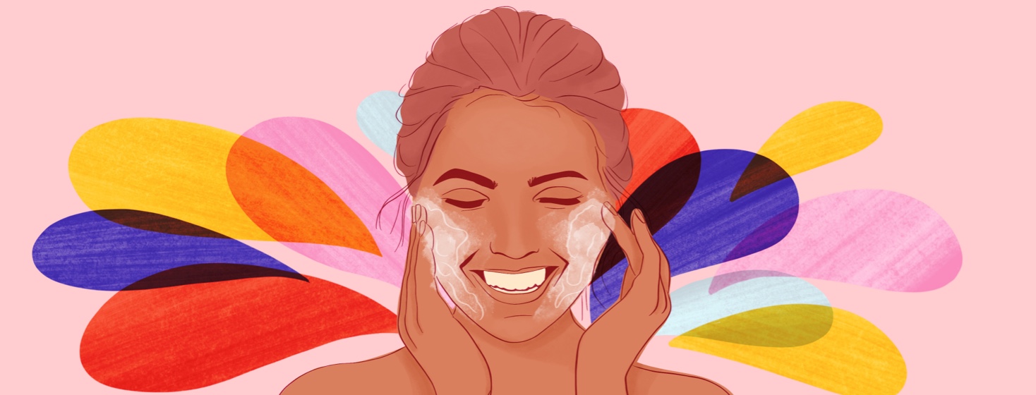 A woman rubs face wash into her cheeks, smiling, multicolored splashes in the background.