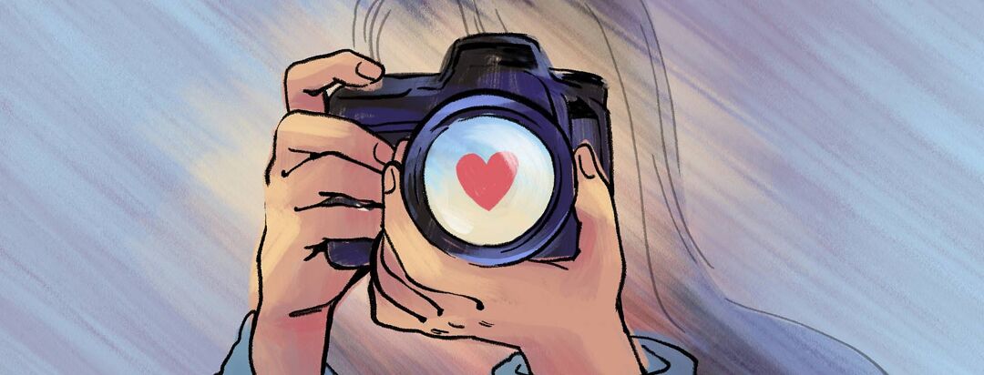 A person holds a camera with a heart shape reflected in the lens.